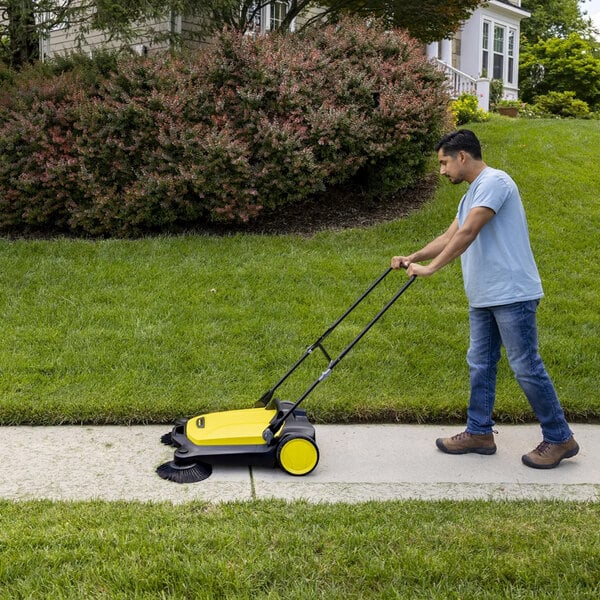 A person is pushing a yellow and black manual sweeper along a sidewalk next to a grassy lawn.
