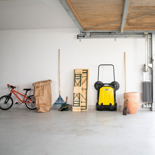 A garage with various items lined up against the wall, including a red bicycle, a large paper bag, a rake, a folded wooden table, a yellow and black manual sweeper, a shovel, and stacked terracotta pots.