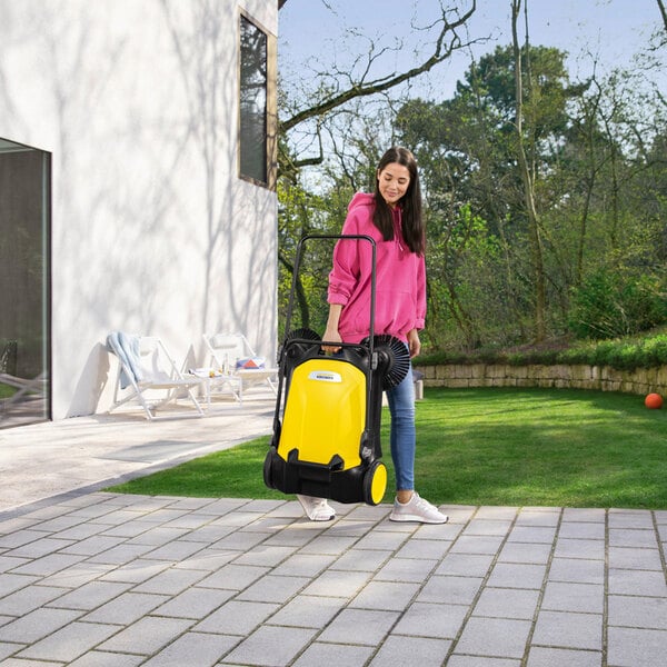 A woman in a pink hoodie is using a yellow and black manual sweeper on a paved outdoor area next to a lawn.