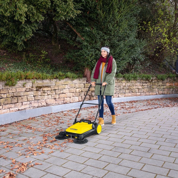 A person is using a yellow manual sweeper to clean leaves from a paved outdoor area.
