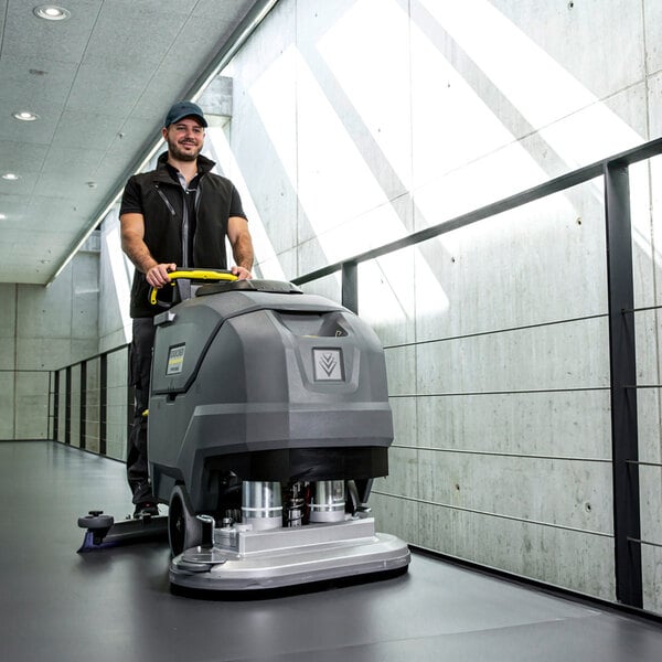 A person is operating a large, gray, walk-behind floor scrubber in a modern hallway with concrete walls and ample natural light.
