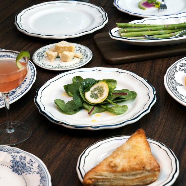 A table set with scalloped white plates featuring a green rim, displaying various foods including a salad with lemon, cheese chunks, asparagus, a pastry, and a cocktail.