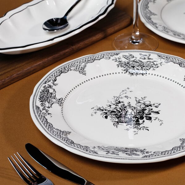 A white round plate with intricate black floral and ornamental patterns, set on a brown table with cutlery and other dishware nearby.