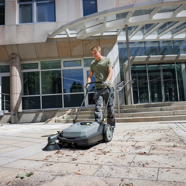 A person is using a large, walk-behind floor sweeper to clean an outdoor paved area in front of a modern building.