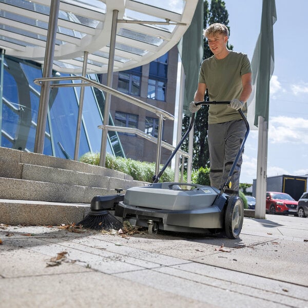 A person is using a large, walk-behind manual floor sweeper to clean debris from an outdoor paved area near steps and a building entrance.