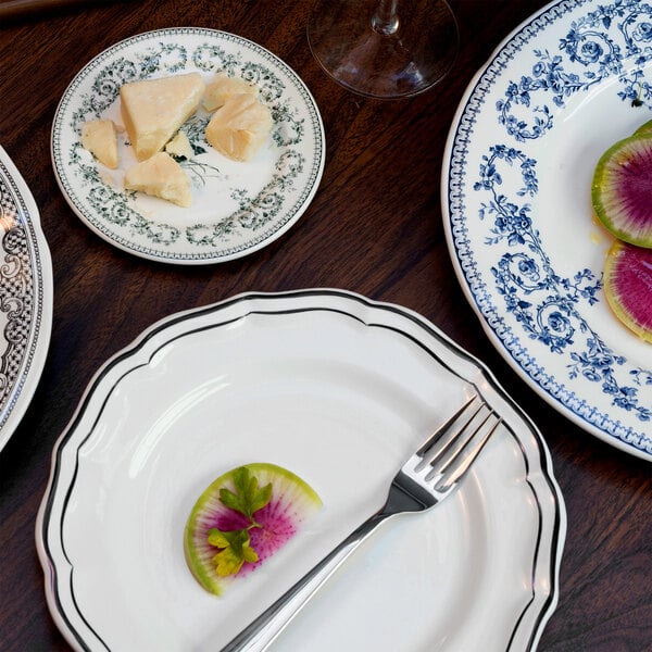 A white scalloped plate with a thin black border, holding a fork and a small garnish, is shown among other decorative plates on a wooden table.