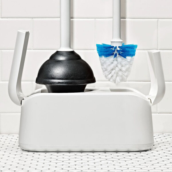 A white caddy holding a black toilet plunger and a white toilet brush with blue bristles, placed on a tiled bathroom floor in front of a white tiled wall.