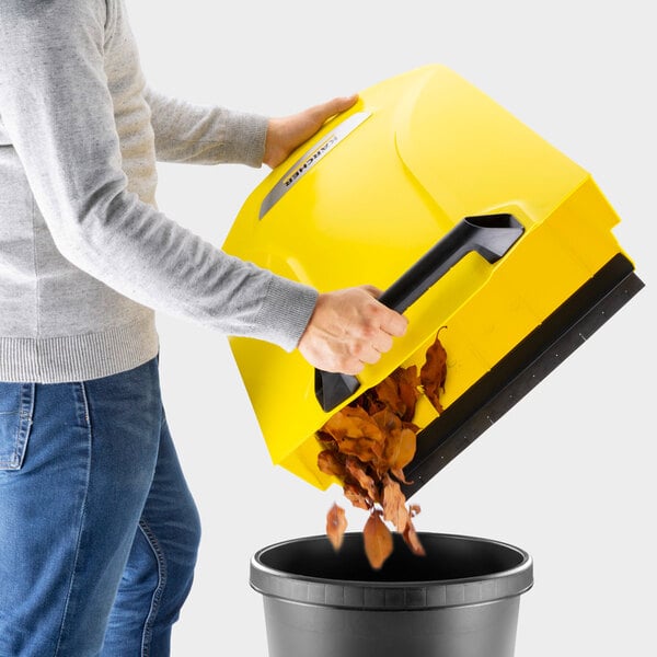 A person is emptying dried leaves from a large yellow container into a black trash bin.