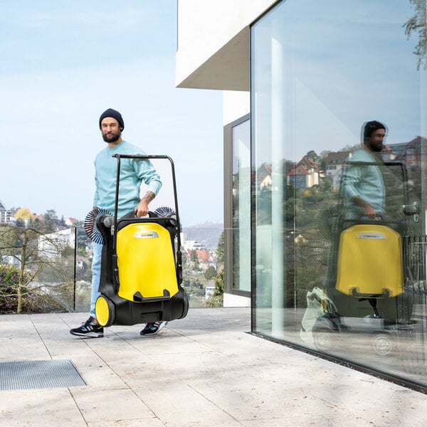A man is standing outdoors on a tiled patio holding a large yellow and black manual sweeper with two side brushes, next to a modern glass building.