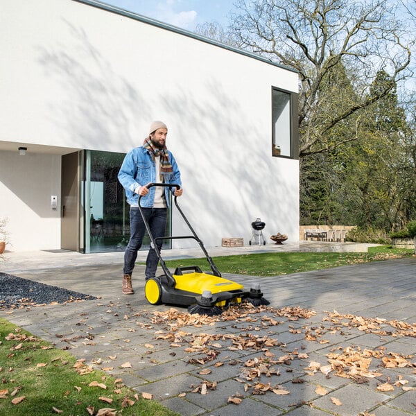 A person is using a yellow and black manual sweeper to clean fallen leaves from a paved walkway outside a modern house.