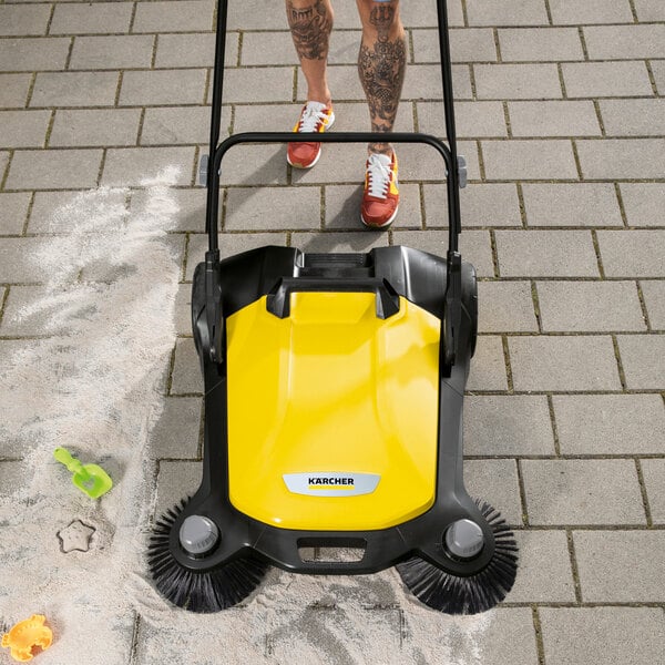 A yellow and black manual sweeper with two rotating brushes is being pushed on a paved surface, cleaning up sand and debris.