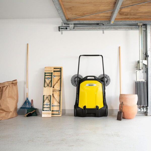 A yellow and black manual sweeper with two side brushes is positioned against a garage wall, surrounded by garden tools, a folded table, and stacked flower pots.