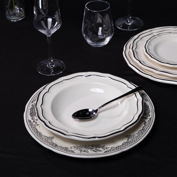 A set table with a white and black scalloped soup plate, a spoon, a matching dinner plate, and glassware on a black tablecloth.