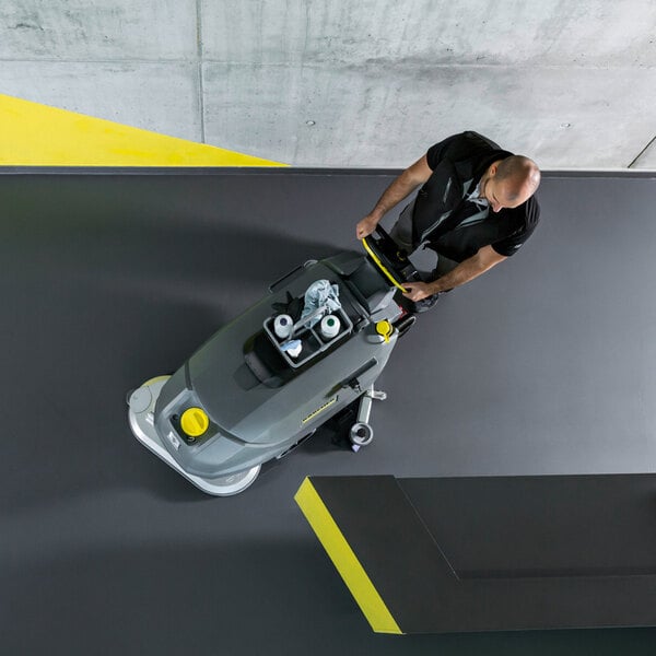 A person is operating a large, gray walk-behind floor scrubber on a smooth, dark floor, viewed from above.