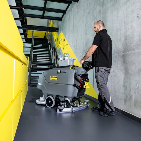 A person is operating a large, gray, walk-behind floor scrubber on a smooth indoor floor near a staircase.