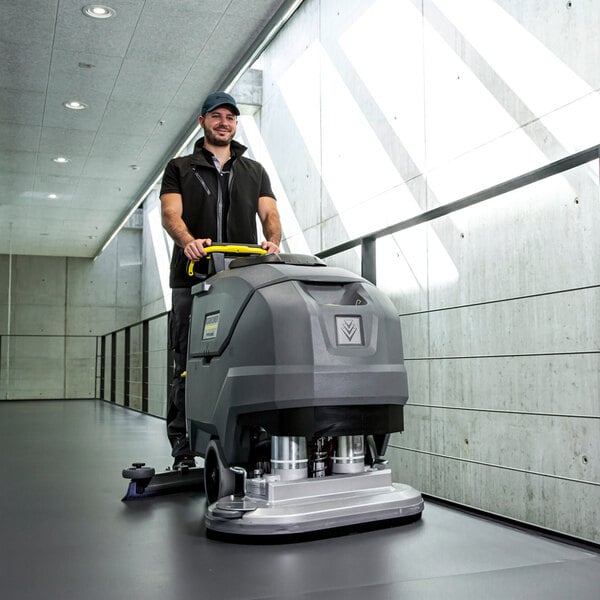 A person is operating a large, gray, walk-behind floor scrubber in a modern hallway with concrete walls and ample natural light.