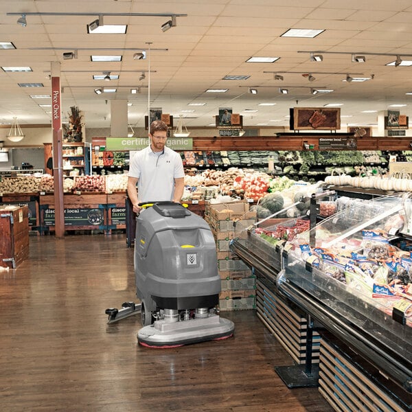 A person is operating a large, gray floor scrubber machine on the wooden floor of a grocery store near the produce section.