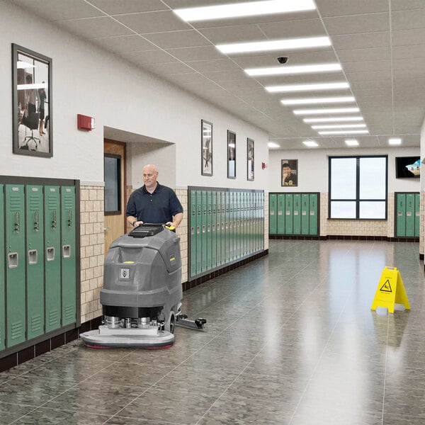 A person is operating a large gray floor scrubber in a hallway lined with green lockers and a caution sign on the floor.