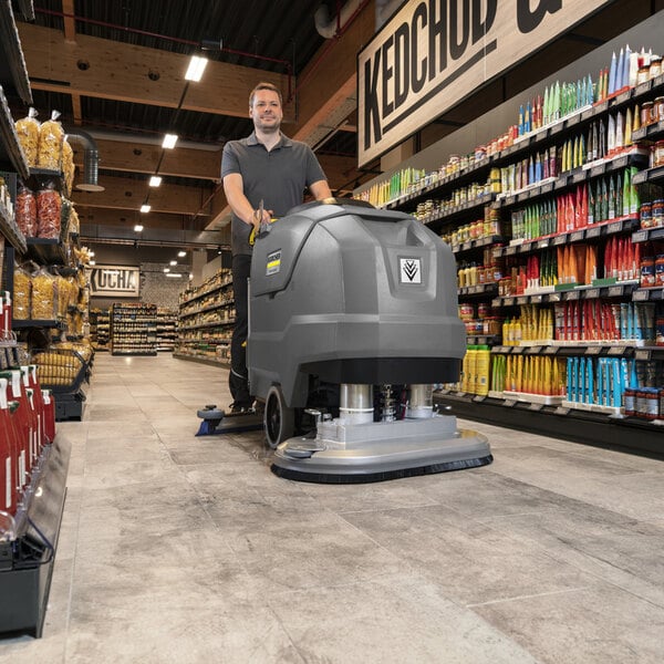 A person is operating a large, gray, walk-behind floor scrubber in a grocery store aisle.