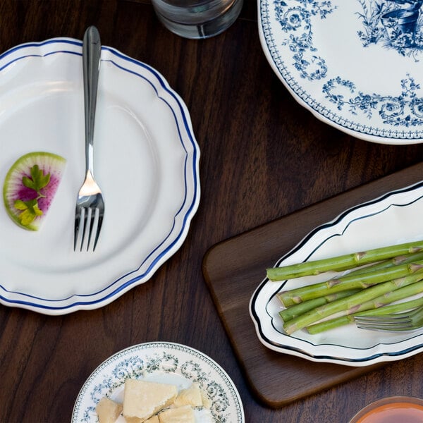 A white scalloped plate with a blue rim, holding a fork and a small garnish, is shown on a wooden table among other plates and food items.