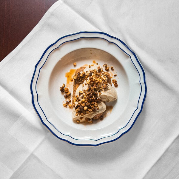 A white scalloped plate with a blue rim containing a dessert topped with a crumbly nut or granola mixture and a drizzle of sauce, placed on a white tablecloth.