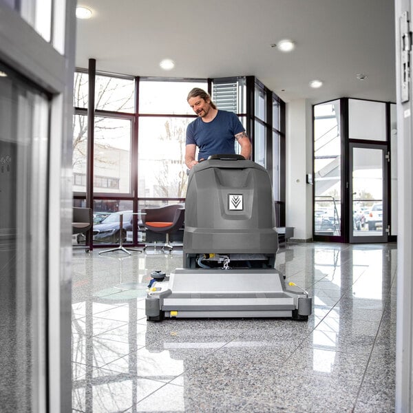 A person is operating a large, gray, walk-behind floor scrubber on a shiny, tiled floor in a modern, well-lit lobby area.
