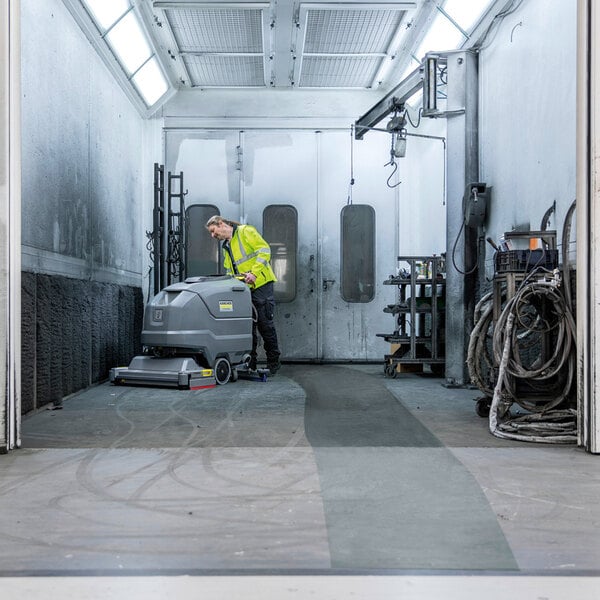 A person in a high-visibility jacket operates a large, gray, walk-behind floor scrubber inside an industrial room with white walls and overhead lighting.