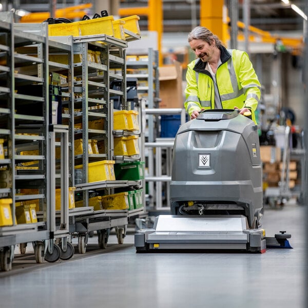 A person in a high-visibility jacket operates a large, gray, walk-behind floor scrubber in an industrial warehouse setting.