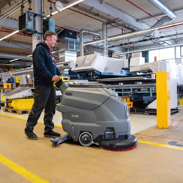 A person is operating a large, gray, walk-behind floor scrubber in an industrial or commercial facility.