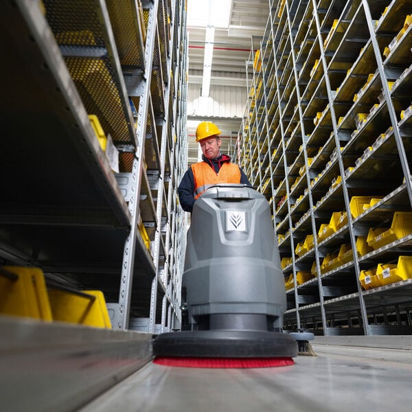 A worker in a hard hat and safety vest operates a large floor scrubber in a warehouse aisle lined with metal shelving and yellow bins.