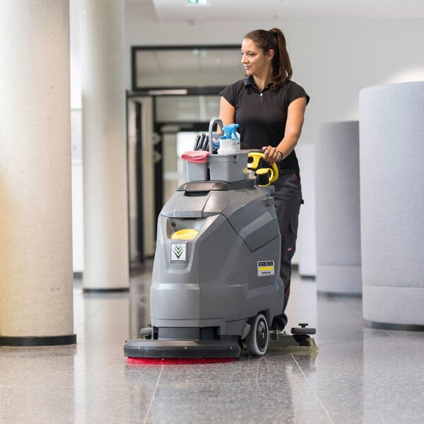 A woman is operating a large, gray, walk-behind floor scrubber on a shiny indoor floor.