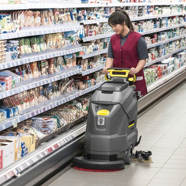 A person is operating a large, gray, walk-behind floor scrubber in a grocery store aisle.