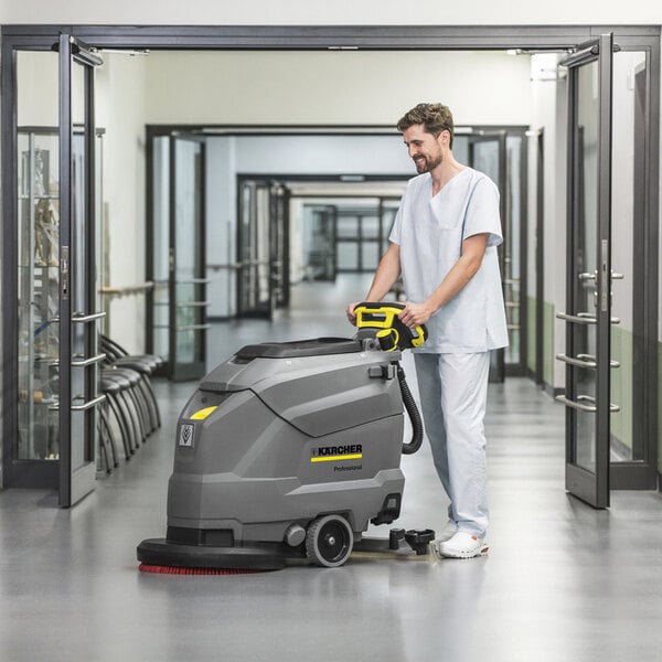 A person in light blue scrubs is operating a large gray and yellow floor scrubber in a clean, modern hallway.