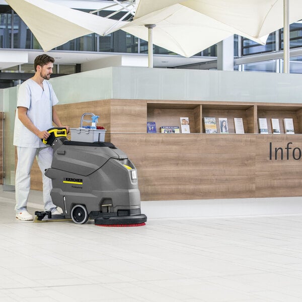 A person is operating a large, gray, walk-behind floor scrubber on a tiled floor in a modern indoor setting.
