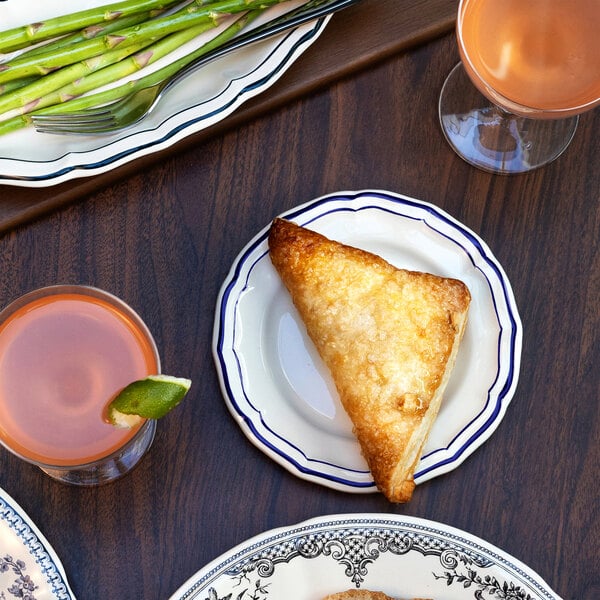 A small white plate with blue scalloped edges holds a golden-brown triangular pastry, surrounded by drinks and a platter of asparagus on a wooden table.