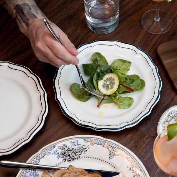 A hand holding a fork is about to eat a small salad with greens and a lemon slice on a white scalloped plate with a green rim, set on a wooden table with other plates and drinks nearby.