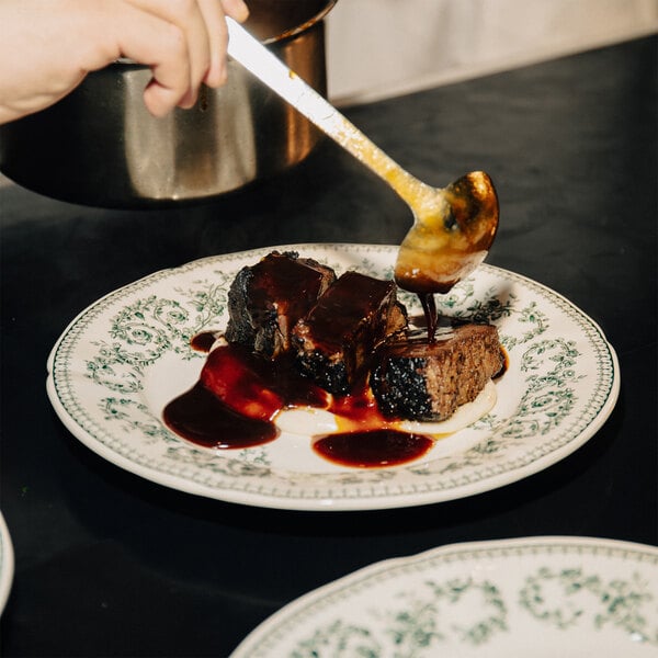 A hand is pouring sauce over two pieces of braised meat on a decorative white plate with a green floral pattern.