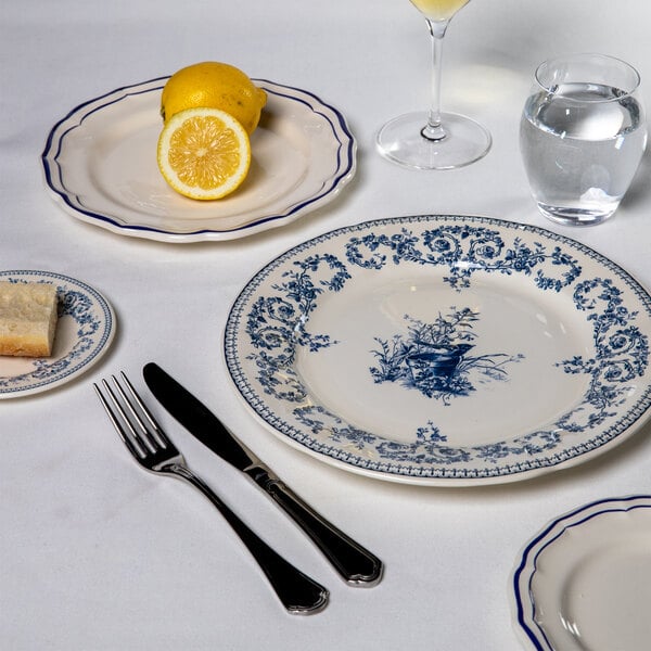 A table setting featuring a white plate with blue floral patterns, accompanied by a fork, knife, a small plate with bread, a plate with a lemon, a glass of water, and a wine glass, all on a white tablecloth.