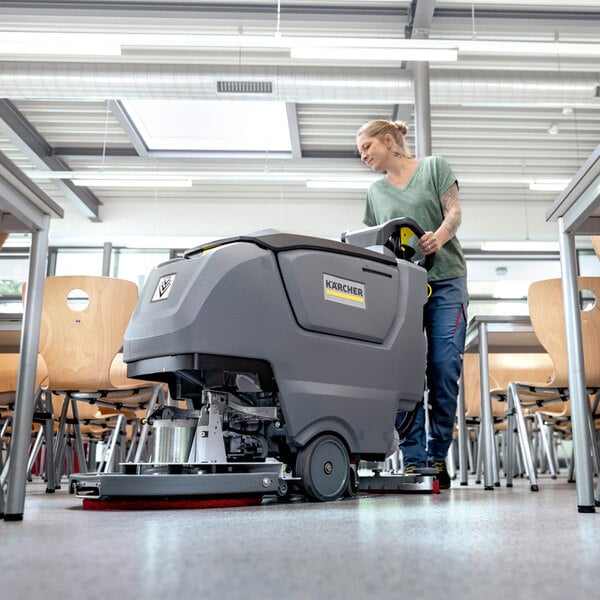 A person is operating a large, gray floor scrubbing machine in a room with wooden chairs and tables.
