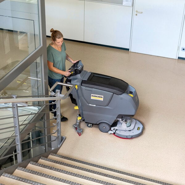 A person is operating a large, gray, walk-behind floor scrubber on a clean indoor floor near a staircase.