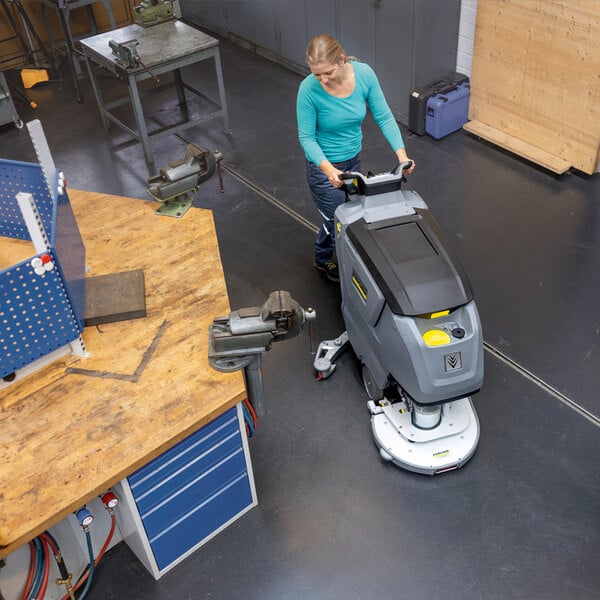 A person is operating a large, gray, walk-behind floor scrubber on a clean, industrial workshop floor.