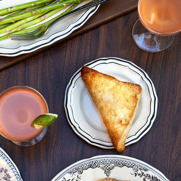 A small scalloped white plate with a black rim, holding a golden-brown triangular pastry, is set on a wooden table alongside drinks and a platter of asparagus.
