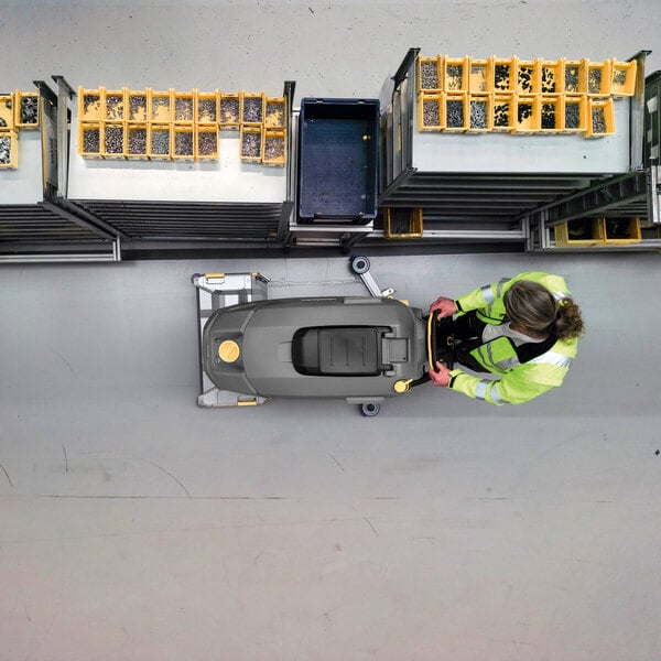 An overhead view of a person operating a large, gray floor cleaning machine in an industrial setting, with organized bins of small parts nearby.