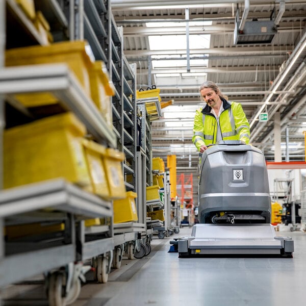 A person in a high-visibility jacket operates a large, gray floor scrubber in an industrial warehouse setting with shelves and yellow bins.