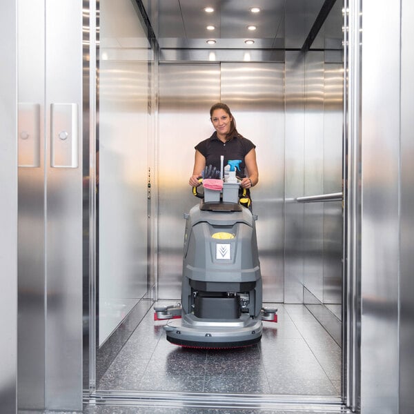 A person is operating a large, gray floor scrubber inside a modern elevator with metallic walls and ceiling lights.