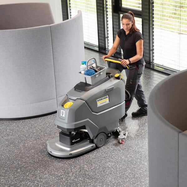 A woman is operating a large, gray, walk-behind floor scrubber on a polished floor in a modern indoor setting.