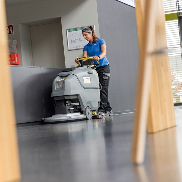 A person is operating a large, gray, walk-behind floor scrubber on a shiny indoor floor.
