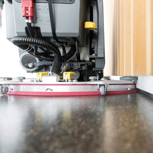 A close-up view of the lower section of a floor cleaning machine operating next to a wall on a polished surface.