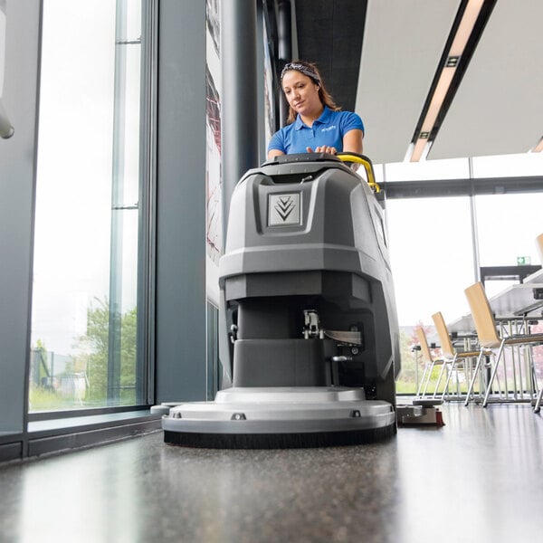 A person is operating a large, gray, walk-behind floor scrubber on a polished floor in a modern indoor space.