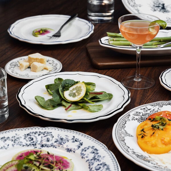 A table set with various scalloped white plates with black trim, featuring salads, cheese, vegetables, and a cocktail.